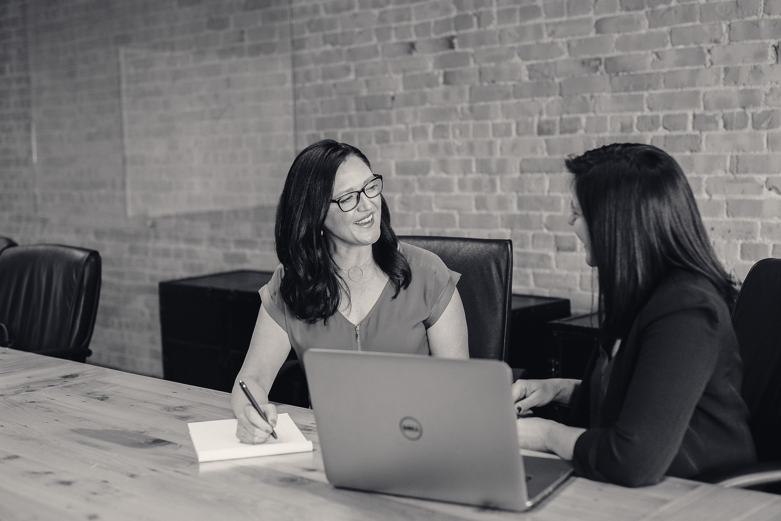 Two colleagues in a working conversation across a table with a laptop and notes